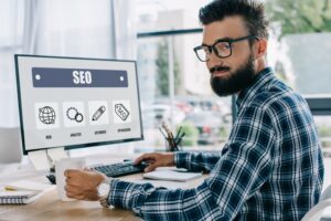 Νέος επιτυχημένος διευθυντής SEO που κάθεται στον χώρο εργασίας με υπολογιστή και κοιτάζει την κάμερα